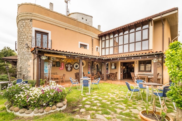 Un edificio con fachada de piedra, un patio cubierto con mesas al aire libre y un jardín con flores.