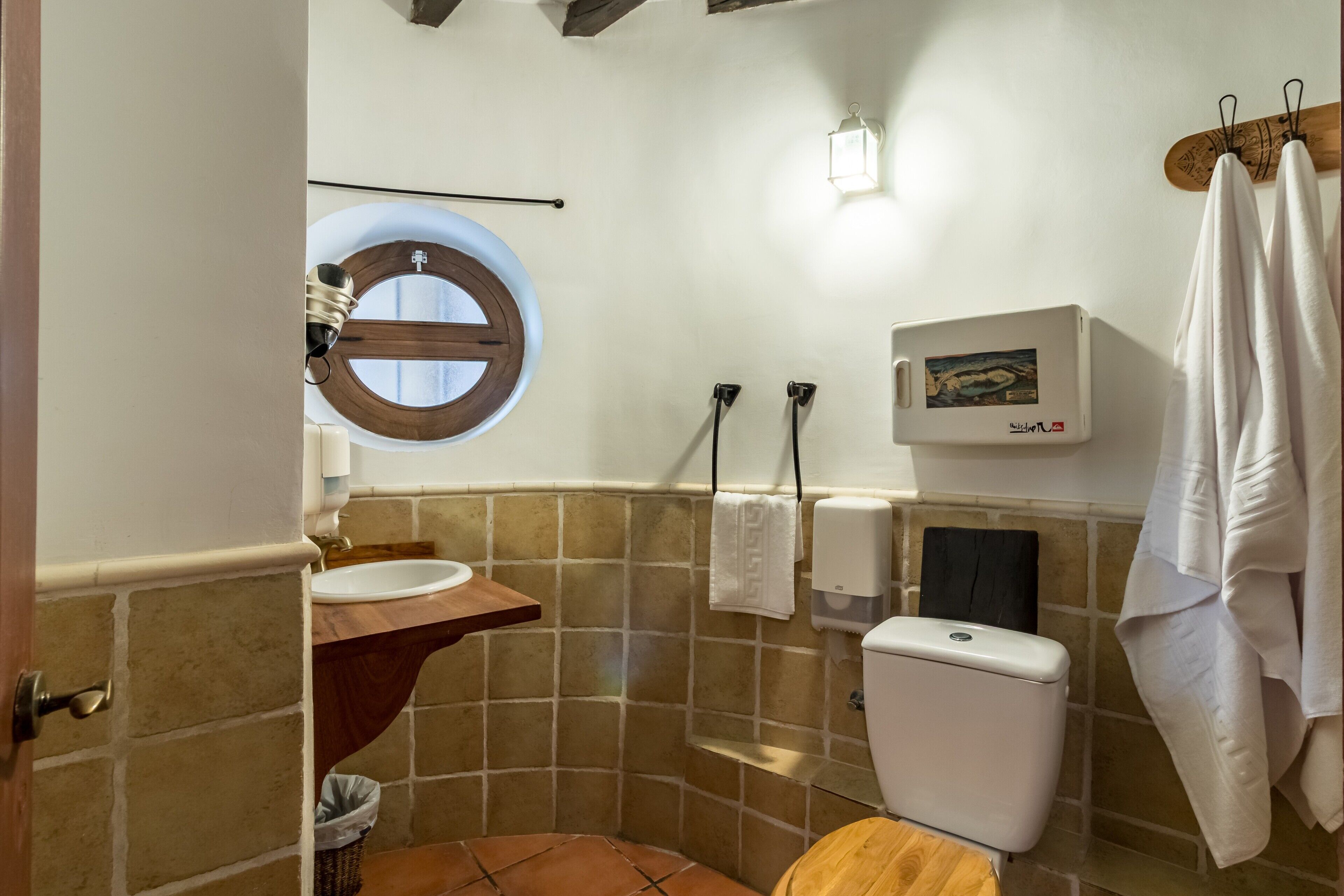 Ein Badezimmer mit Toilette aus Holz, einem Waschbecken, einem Handtuchhalter, einem Seifenspender und einem Fenster mit Holzrahmen.