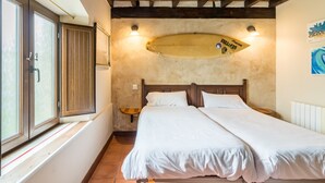 A bedroom with a bed, wooden headboard, a surfboard decoration, a window with curtains, and a wall-mounted light.