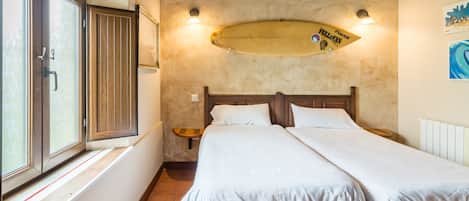 A bedroom with a bed, wooden headboard, a surfboard decoration, a window with curtains, and a wall-mounted light.