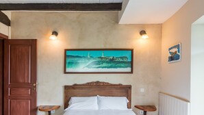 A bedroom with a wooden headboard, a bed with white linens, two bedside tables, a painting of a coastal scene, and a small artwork on the wall.