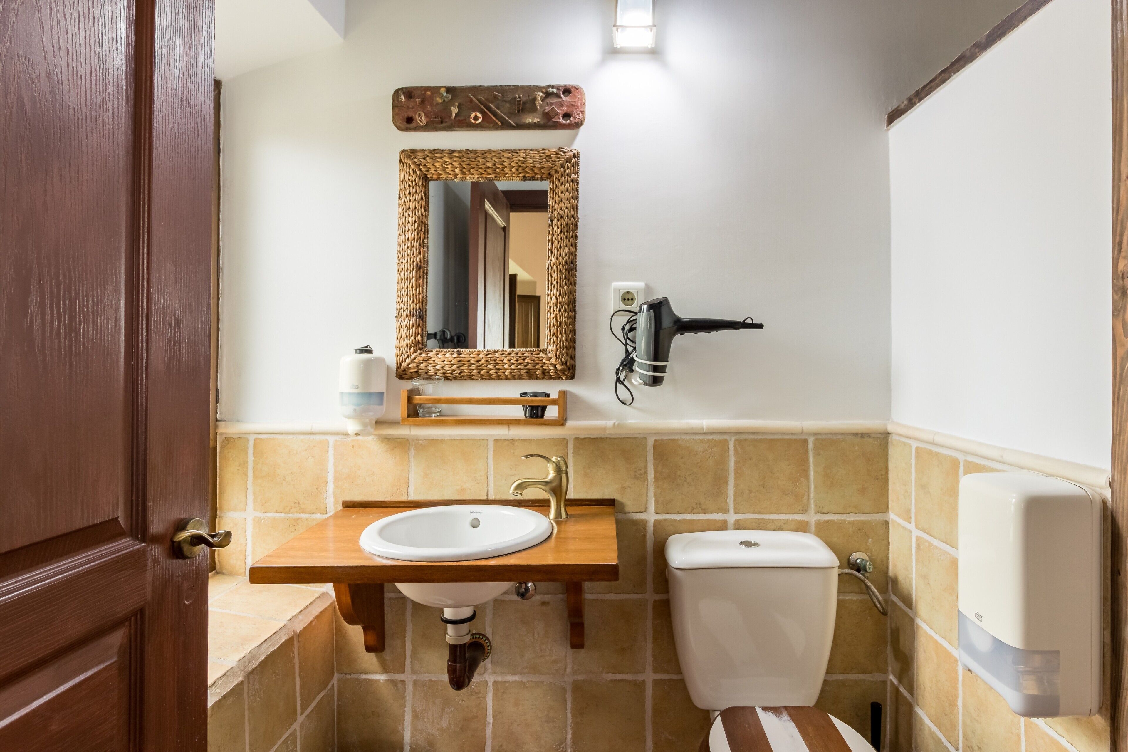 A bathroom with a wooden vanity, a white sink, a toilet, a wall-mounted hairdryer, and a mirror with a woven frame.