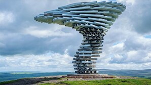 A large, abstract sculpture made of cylindrical metal pieces, set on a grassy hill with a backdrop of cloudy sky and distant landscape. - James Lodge Rishton (Rishton)