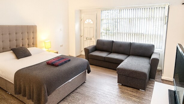 A bedroom with a bed, a grey sofa, a television, and a window with blinds. - James Lodge Rishton (Rishton)