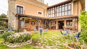Un edificio con fachada de piedra, un patio cubierto con mesas al aire libre y un jardín con flores.