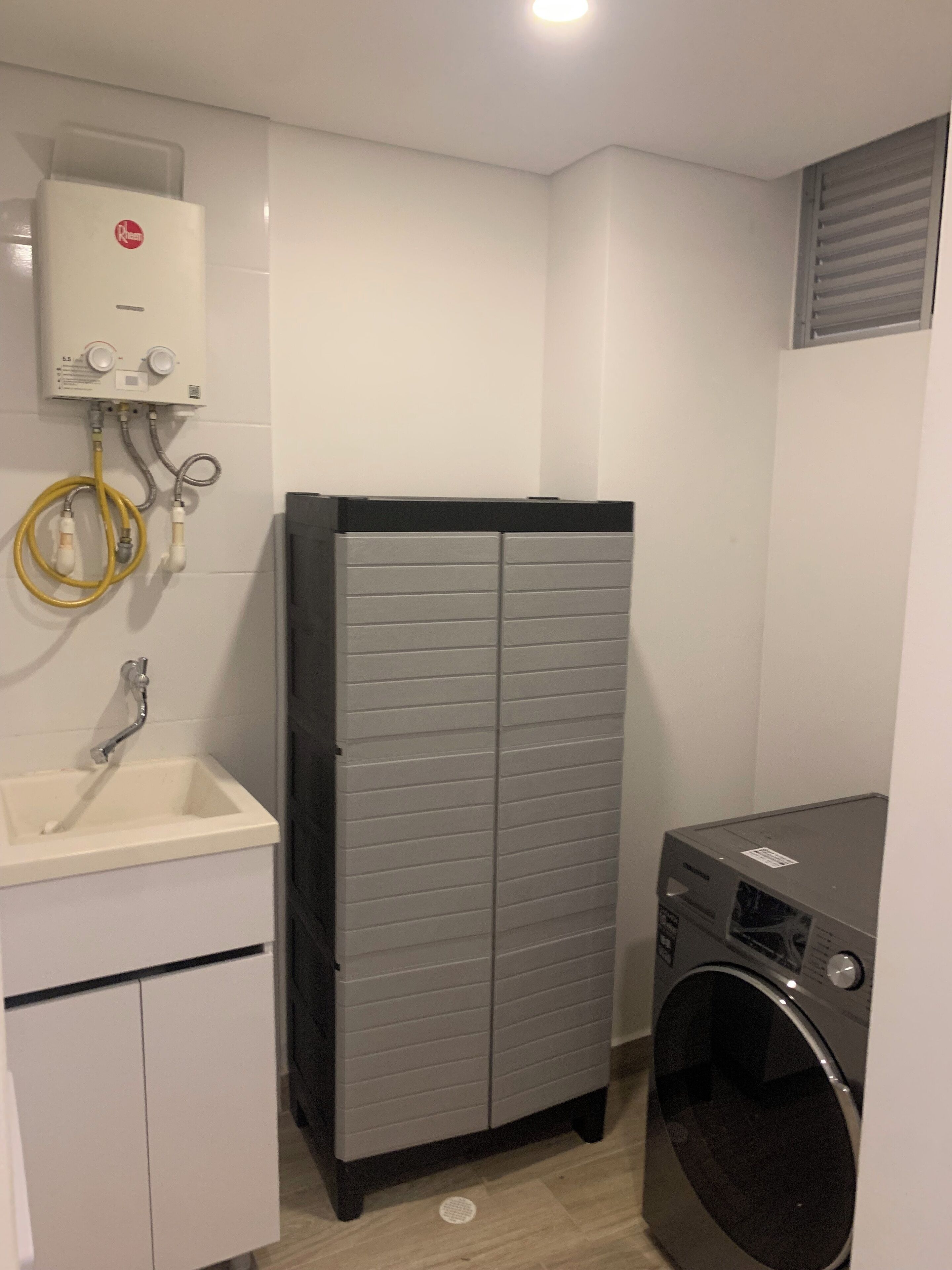 A compact bathroom with a white sink, a grey utility cabinet, a washing machine, and a water heater mounted on the wall.