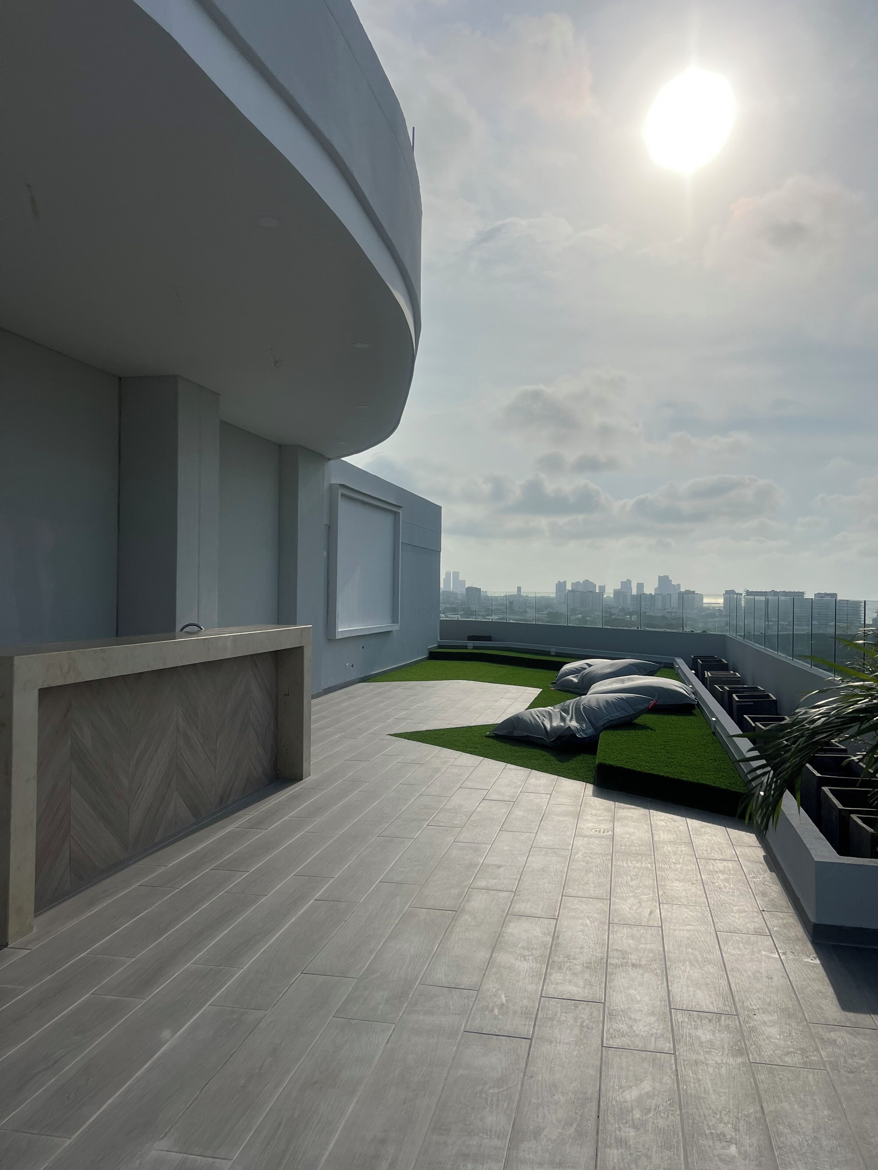 A rooftop terrace with a modern design, featuring a green artificial turf area, a seating arrangement with cushions, and a view of the city skyline.