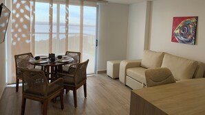 En moderne stue med spiseplads, med en sofa, fletstole, et spisebord med tallerkener og kopper, og et farverigt kunstværk på væggen.