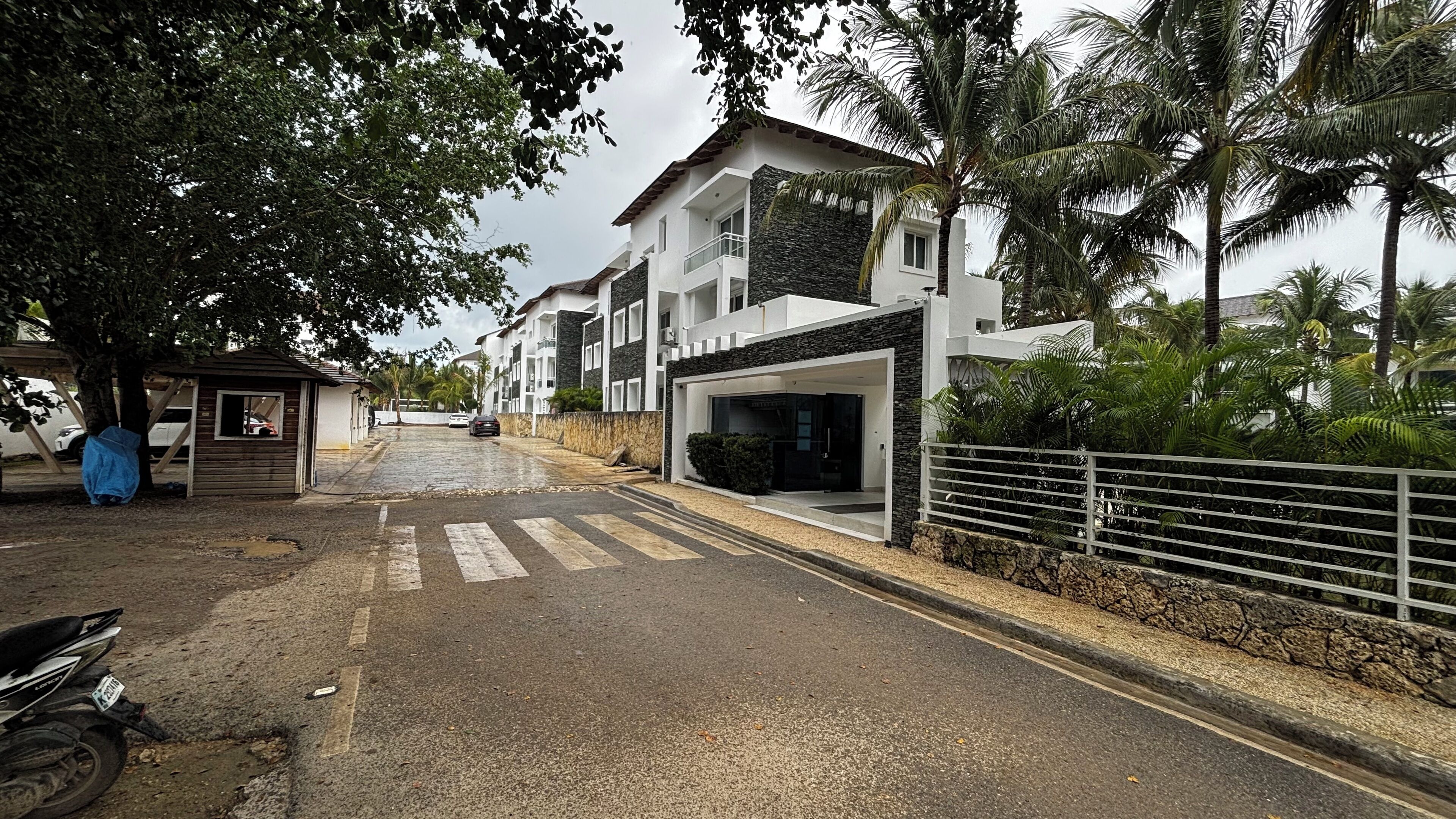 A modern residential street with a paved road, a covered parking area, and a building with a stone facade and balconies.