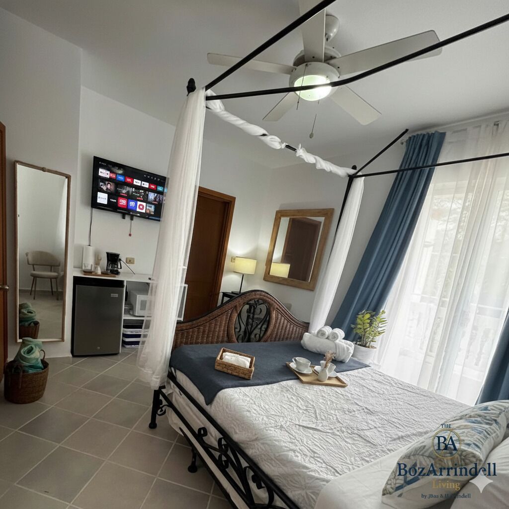 Une chambre à coucher avec un lit à baldaquin, un ventilateur de plafond, un téléviseur fixé au mur et un bureau équipé d’un ordinateur et d’une lampe.