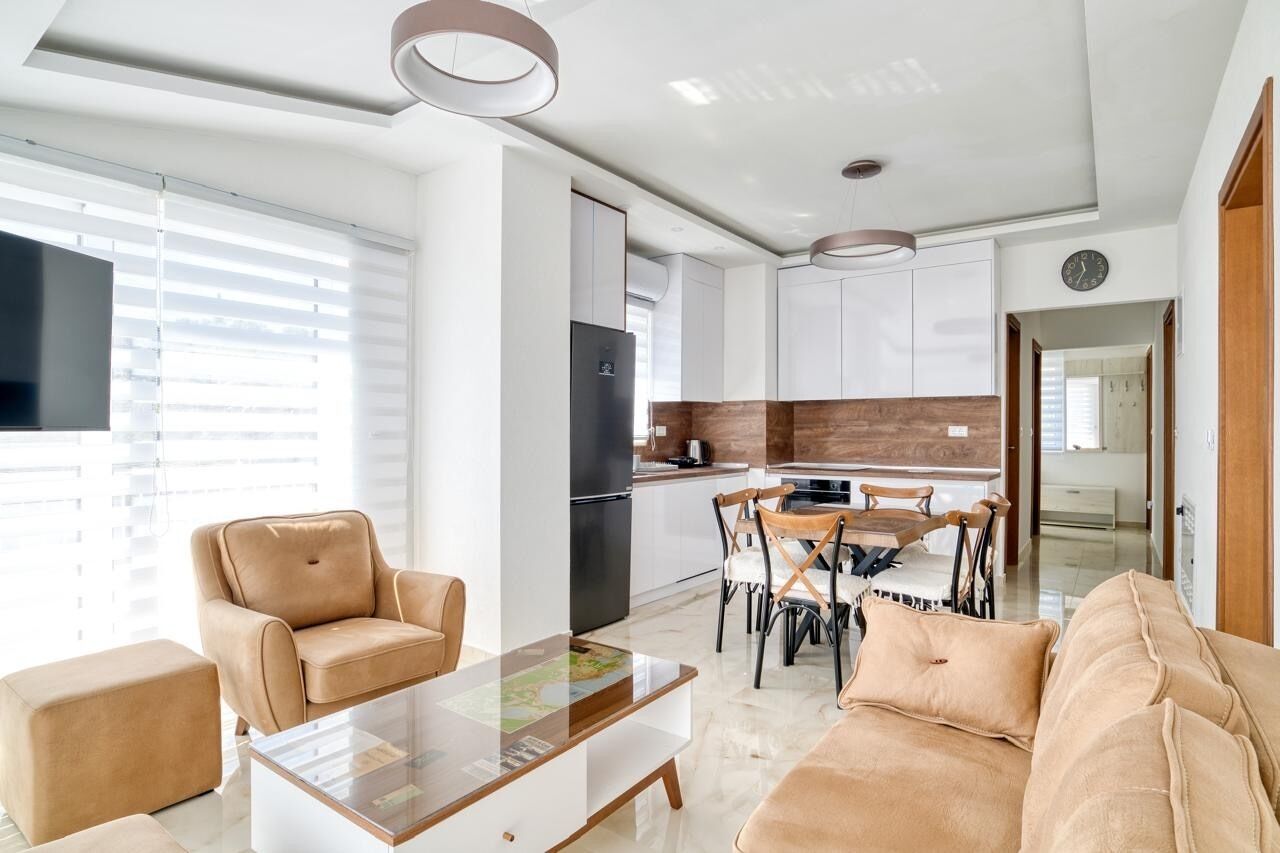 A modern living room with a sofa, coffee table, and chairs. There is a kitchen area with cabinets and a refrigerator.
