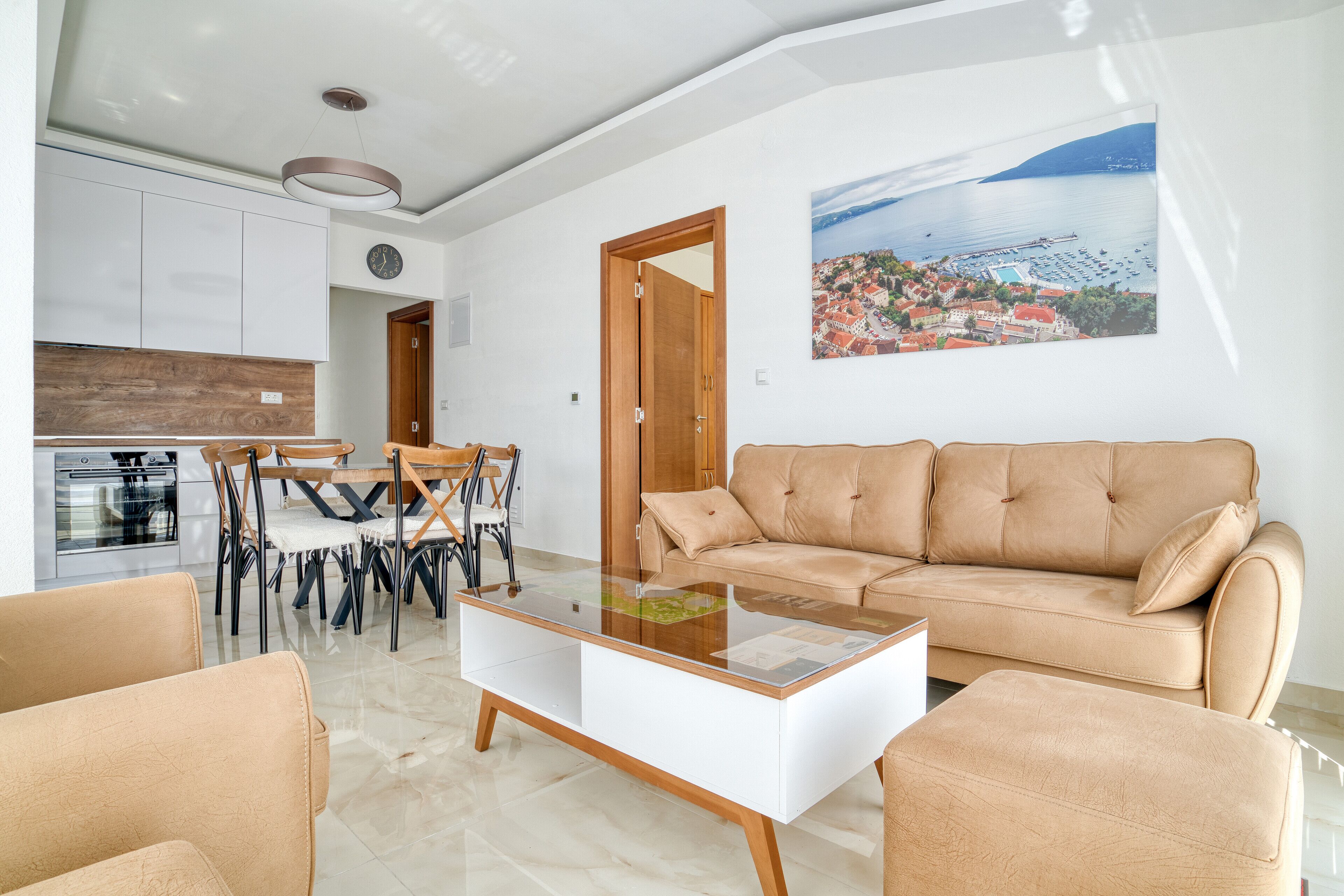 Un salón moderno con un sofá color beige, una mesa de centro de vidrio y sillones a juego. Se ve un comedor con una mesa y sillas de madera al fondo.