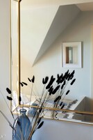 A room with a large mirror reflecting a vase with tall grasses and a framed artwork on the wall.