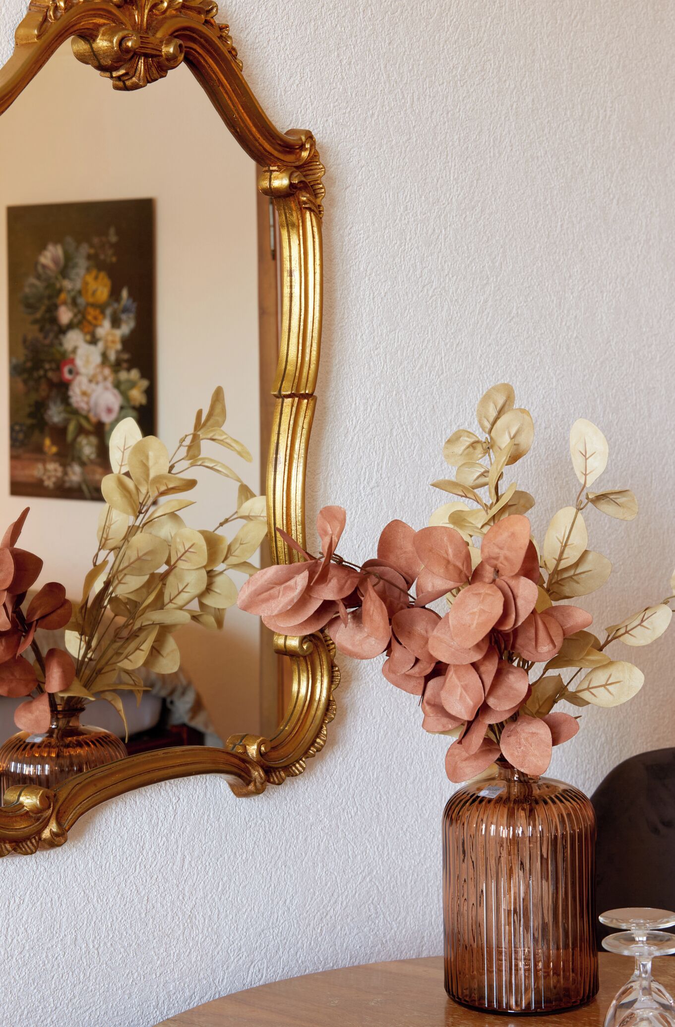 Un miroir avec un cadre doré orné, un vase contenant des feuilles roses et vertes, et un tableau en arrière-plan.