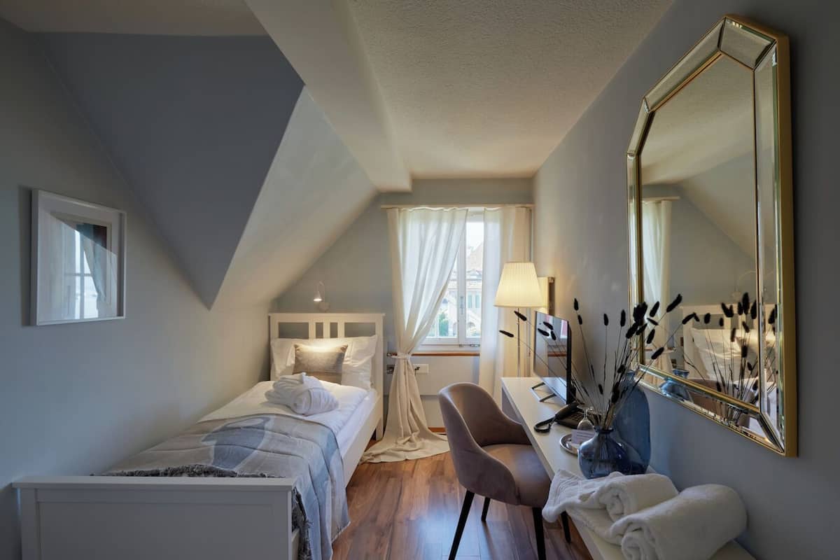 A bedroom with a bed, a desk, a chair, a mirror, and a window with curtains.