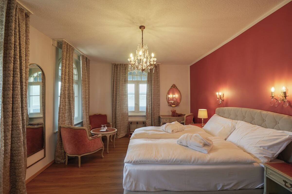 A hotel room with a large bed, a sitting area with a chair and a table, a chandelier, and a mirror.