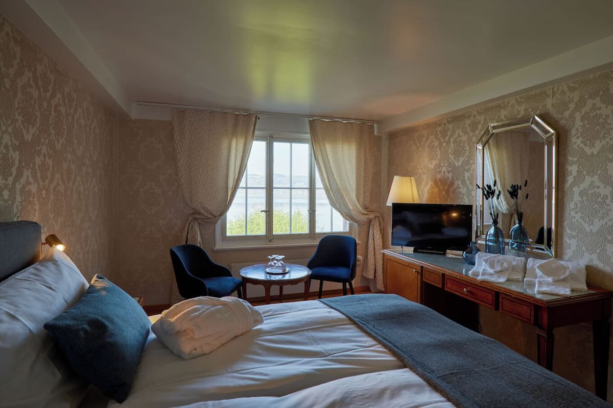A hotel room with a bed, a desk with a TV, a chair, a small table, and a window with curtains.