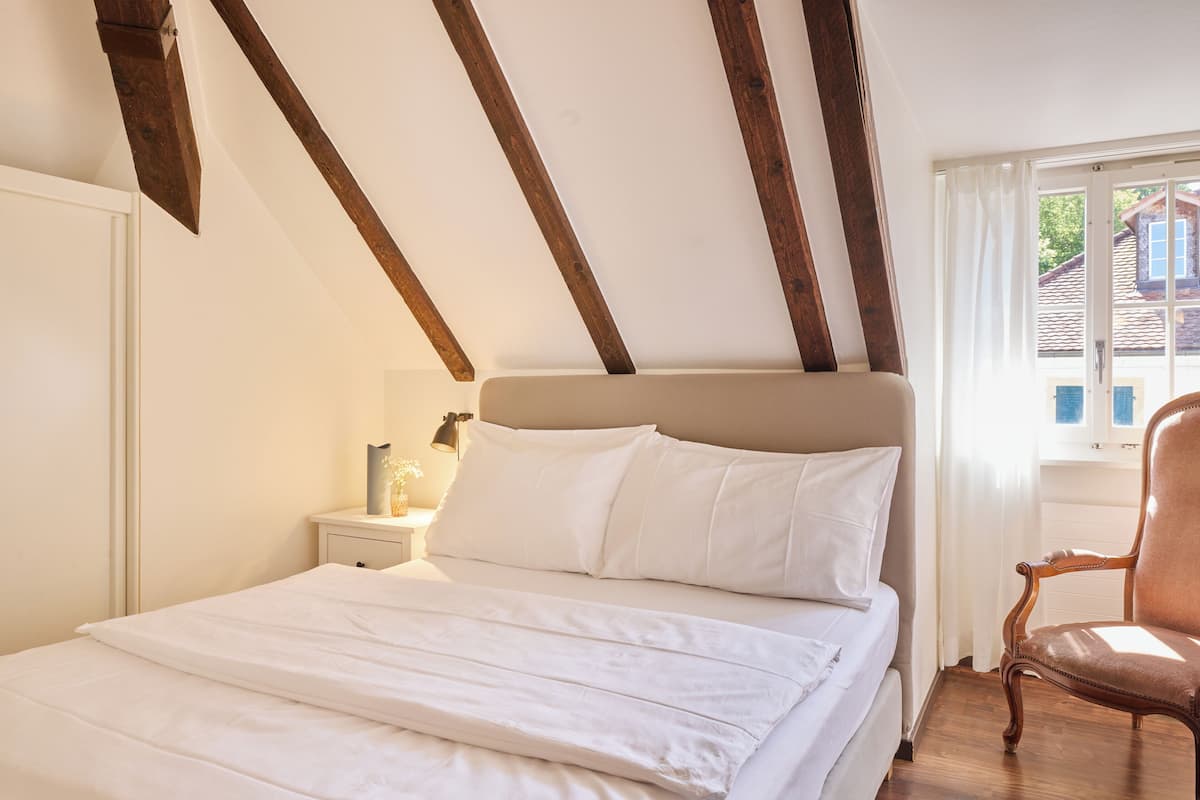 A bedroom with a bed, wooden beams, a nightstand, and a chair.