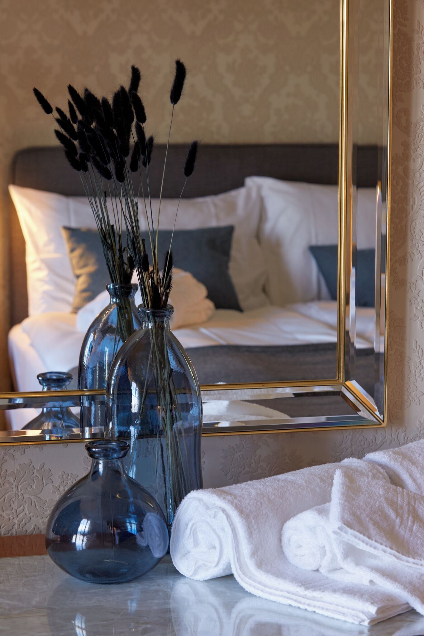 Une salle de bain avec un miroir, deux vases en verre contenant des plantes séchées et une serviette blanche posée sur un comptoir en marbre.