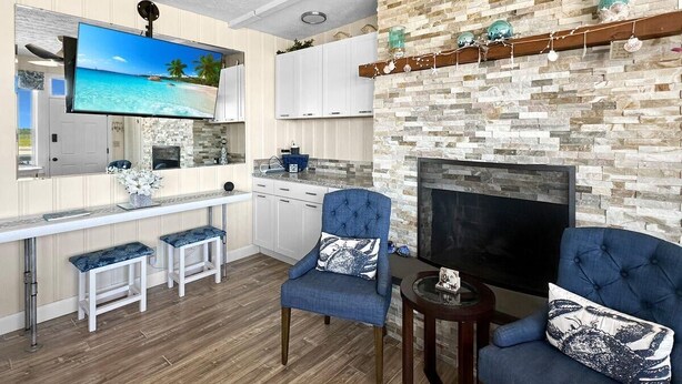 A modern living room with a stone fireplace, a flat-screen TV, and a kitchen area with white cabinets and a wooden countertop. - Admiral's Quarters (Wells)