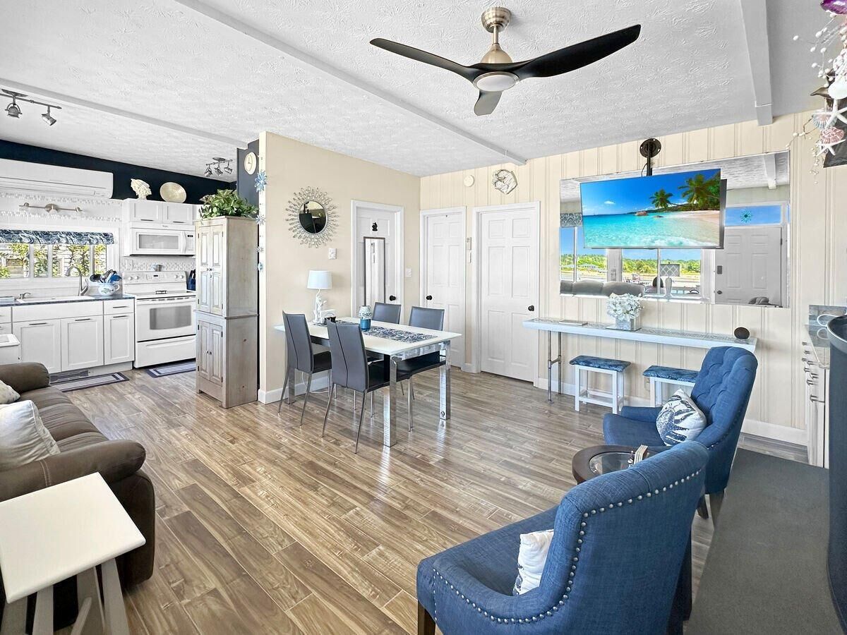 A modern kitchen with white cabinets, a dining area with a table and chairs, and a living room with a blue armchair and a sofa.