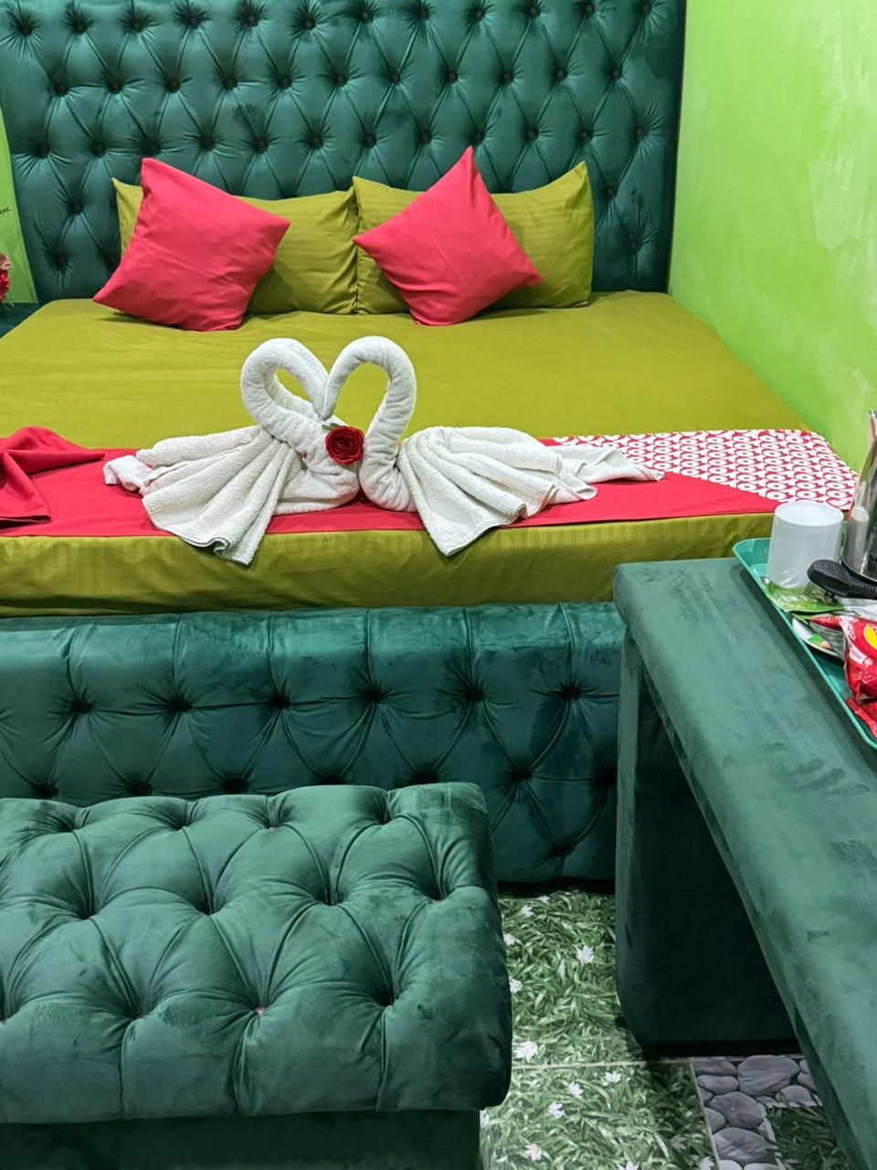 Una cama con cabecera verde, almohadas rojas y amarillas, y una toalla con forma de cisne. Hay un reposapiés verde y una mesita con una bandeja y una taza.