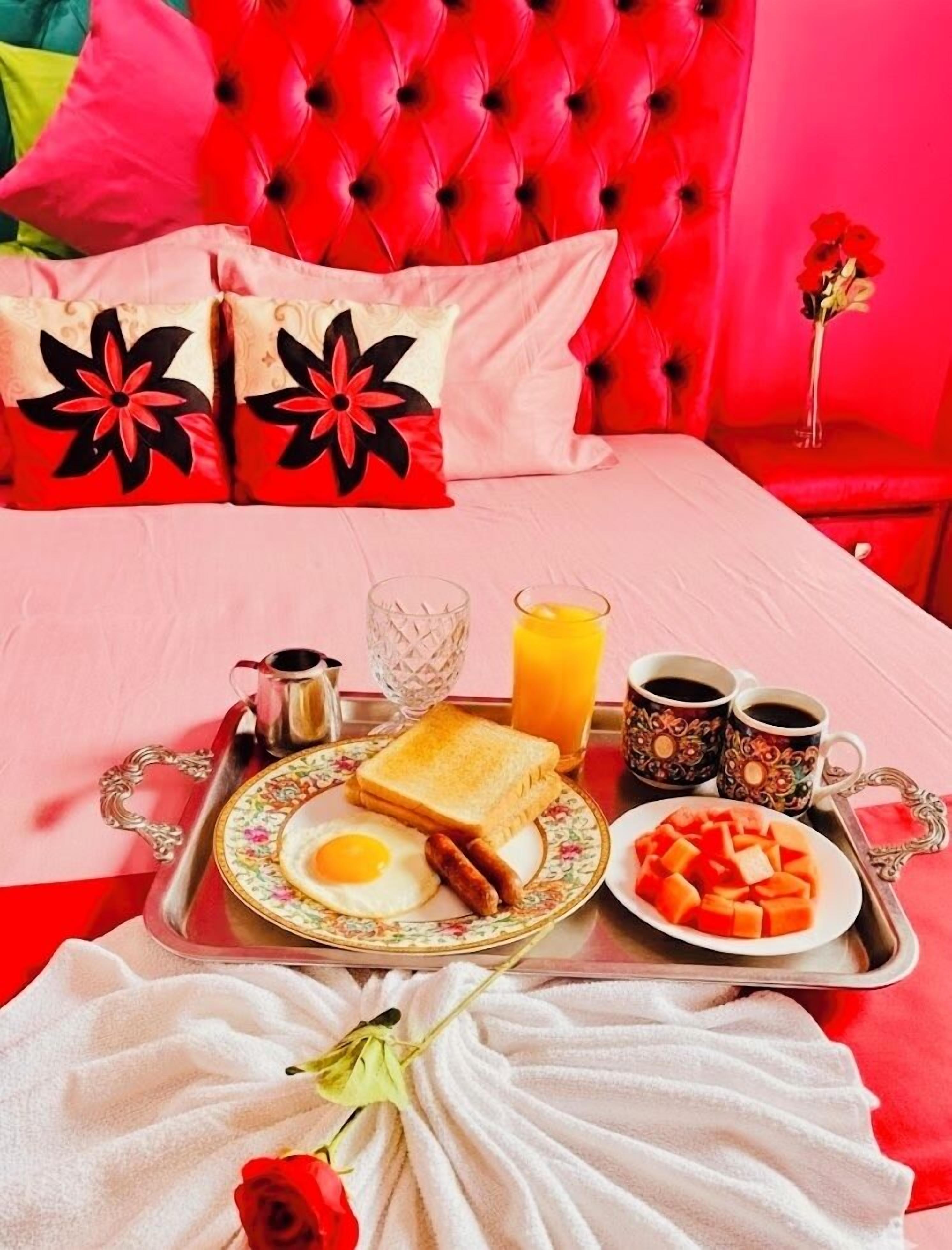 Bandeja de desayuno ordenada con tostadas, huevos, salchichas y fruta, sobre una cama con cabecera roja y almohadas estampadas con motivos florales.