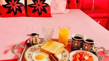 Bandeja de desayuno ordenada con tostadas, huevos, salchichas y fruta, sobre una cama con cabecera roja y almohadas estampadas con motivos florales.
