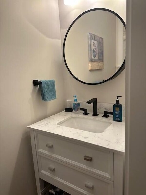 A bathroom with a white marble sink, a black faucet, a round mirror, and a towel rack with a blue towel. - Seaglass Village 500 - Seaglass Village Gem (Wells)