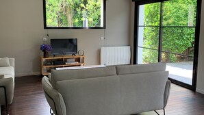 Un salon moderne avec un grand canapé gris, un plancher en bois, un téléviseur posé sur un meuble en bois et une fenêtre donnant sur un paysage verdoyant.