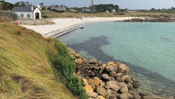 Un paysage côtier avec une plage de sable, une digue rocheuse et un phare au loin.