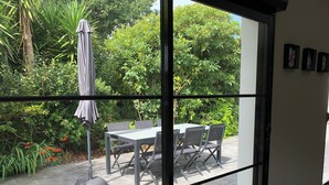 Une chambre moderne dotée d’une grande porte-fenêtre coulissante qui donne sur une terrasse extérieure avec une table et des chaises, le tout entouré d’une végétation luxuriante.