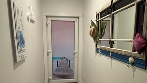 Ein Flur mit Holzboden, einer Tür, die Blick auf einen Strand bietet, einem wandmontierten Spiegel und einem kleinen Fenster mit Pflanze und Hut.