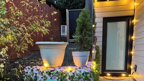Eine Terrasse mit blauem Korbstuhl, zwei großen Pflanzgefäßen und Lichterketten.