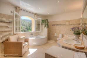 Ein Badezimmer mit freistehender Badewanne, zwei Waschbecken, einer Korbsessel-Couch und einem großen Fenster mit Blick auf Grünflächen.