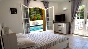 Een slaapkamer met een bed, een televisie, een schuifdeur naar een zwembad en uitzicht op groen.