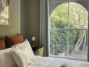 A bedroom with a large arched window, a bed with white and brown pillows, a bedside table with a lamp, and a view of greenery outside. - Victorian 2BR Islington Flat|Garden, Near Upper St - Pass the Keys (London)