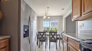 A kitchen with wooden cabinets, a stainless steel refrigerator, a dining table with chairs, and a window with a view of houses. - Grey Moon Escape In Bloomington (Bloomington)
