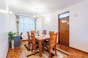 Ein Essbereich mit einem Holztisch, gedeckt für sechs Personen, umgeben von Stühlen. Links befindet sich ein Blumentopf, auf dem Boden ein Teppich und rechts eine Tür.