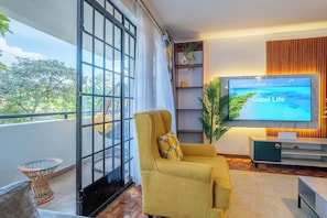 Ein modernes Wohnzimmer mit einem gelben Sofa, einem Fernseher, der 'Good Life' zeigt, und einem Balkon mit Blick auf Grünflächen.