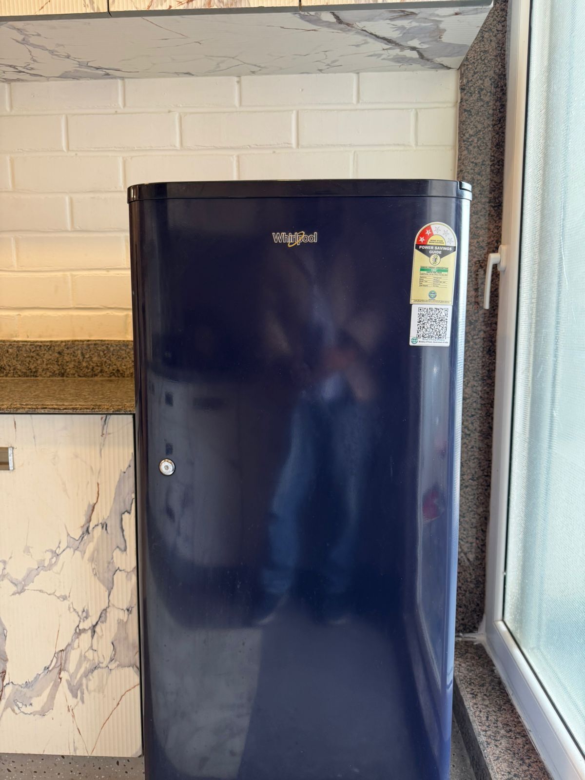 A black Whirlpool refrigerator with a power savings guide sticker, placed in a kitchen with white brick walls and a granite countertop.