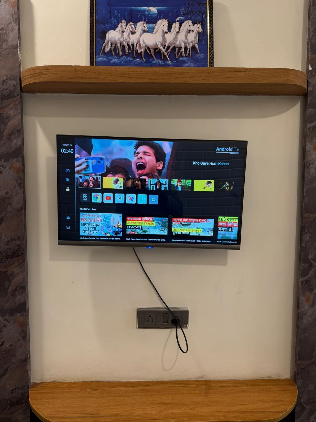 Een aan de muur bevestigde Android TV toont een video met meerdere app-icoontjes en een YouTube Live kanaal.