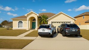 A two-story house with a garage, two parked SUVs, and a palm tree in the front yard. - 5BR Disney Retreat | Private Pool, Spa & Game Room — Sleeps 14 (Kissimmee)