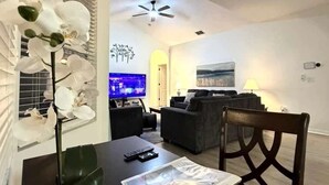 A modern living room with a dining table, a sofa, a TV, and a ceiling fan. - 5BR Disney Retreat | Private Pool, Spa & Game Room — Sleeps 14 (Kissimmee)