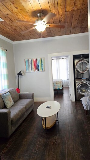 A living room with a ceiling fan, a couch, a coffee table, a painting, a lamp, and a washing machine. - Adorable 1-bedroom house w/private entrance, beach 2 blks in charming Galveston (Galveston)