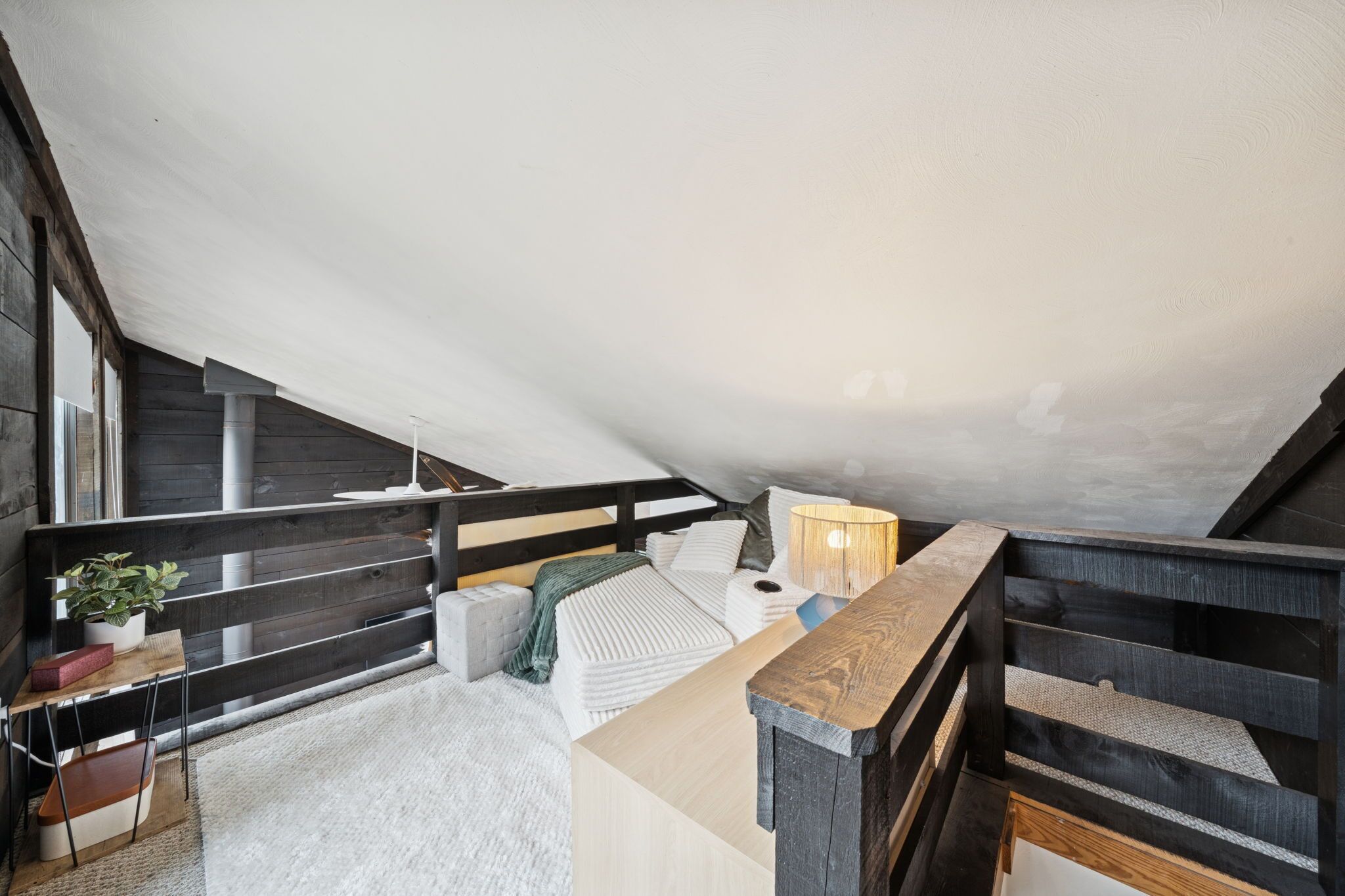 A loft bedroom with a sloped ceiling, wooden beams, a bed with white bedding, a small table with a plant, and a lamp.