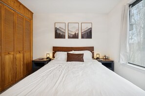Un dormitorio con un armario de madera, una cama con ropa de cama blanca, un almohón marrón y tres cuadros enmarcados con paisajes de montaña en la pared.