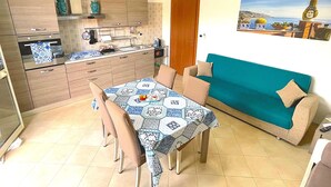 A compact kitchen with modern appliances, a dining table with chairs, and a teal sofa against a wall with a landscape painting. - The Star of the South - Holiday Home. Apartment at the foot of Etna (Mascali)