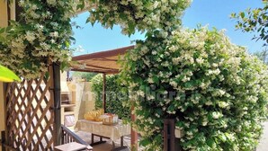 Un espacio de estar al aire libre cubierto, con una mesa y sillas, rodeado de abundante follaje y plantas con flores.