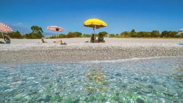 Una playa con aguas poco profundas y transparentes, piedras y gente relajándose bajo sombrillas.